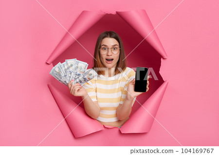 Indoor shot of excited woman wearing striped shirt looking through breakthrough of pink background, standing with smart phone with blank scree and dollar banknotes, cashback. Indoor shot of excited woman wearing striped shirt looking through breakthrough of pink background, standing with smart phone with blank scree and dollar banknotes, cashback. 104169767