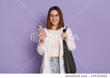 Image of adorable woman with black bag wearing white shirt and jeans posing isolated over purple background, using mobile phone, checking new app, looking smiling at camera. Image of adorable woman with black bag wearing white shirt and jeans posing isolated over purple background, using mobile phone, checking new app, looking smiling at camera. 104169802