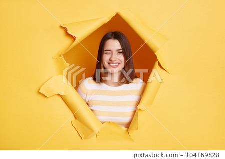 Indoor shot of playful young adult woman stands in torn paper hole, looking through breakthrough of yellow background and winking, expressing positive emotions and happiness. 104169828
