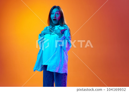 Shock and surprise. Portrait of beautiful young girl in casual clothes posing against gradient orange studio background in neon light Shock and surprise. Portrait of beautiful young girl in casual clothes posing against gradient orange studio background in neon light 104169932