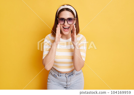 Image of positive delighted dark haired woman wearing striped t shirt and jeans posing isolated over yellow background, standing with hands near mouth and screaming, making an announce. 104169959