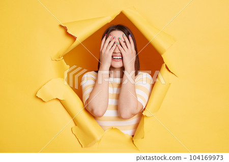 Horizontal shot of unknown anonymous woman stands in torn paper hole, posing and covering eyes with palms, looking through breakthrough of yellow background. Horizontal shot of unknown anonymous woman stands in torn paper hole, posing and covering eyes with palms, looking through breakthrough of yellow background. 104169973