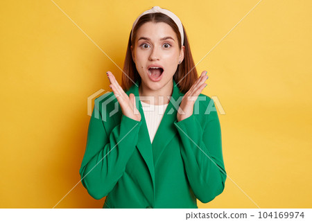 Portrait of extremely surprised woman wearing green jacket and hair band, raised arms, keeps mouth widely opened, being very shocked, posing isolated over yellow background. Portrait of extremely surprised woman wearing green jacket and hair band, raised arms, keeps mouth widely opened, being very shocked, posing isolated over yellow background. 104169974