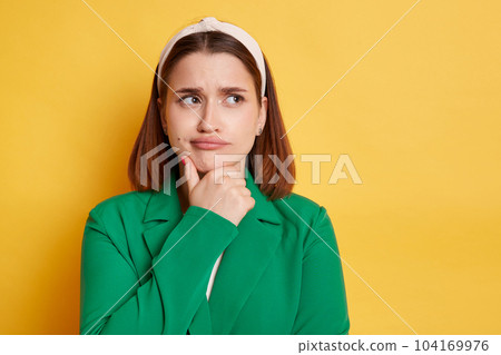 Portrait of confused attractive young woman wearing stylish green jacket looking side with pensive expression, holding chin, thinking, posing isolated over yellow background. 104169976