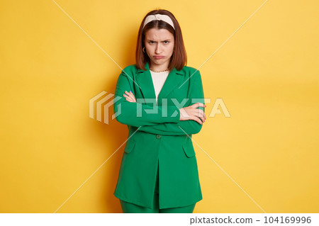 Indoor shot of sad upset dark haired Caucasian young woman wearing stylish green jacket posing isolated over yellow background, standing with folded hands and pout lips, being offended. Indoor shot of sad upset dark haired Caucasian young woman wearing stylish green jacket posing isolated over yellow background, standing with folded hands and pout lips, being offended. 104169996