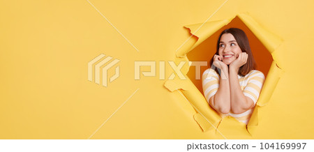 Smiling delighted woman wearing striped t shirt, expreressing positve emotions, copy space for advertisement or promotional text, posing standing through yellow paper torn hole. 104169997