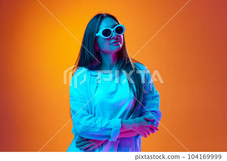 Portrait of young woman in casual clothes posing in trendy sunglasses against gradient orange studio background in neon light Portrait of young woman in casual clothes posing in trendy sunglasses against gradient orange studio background in neon light 104169999