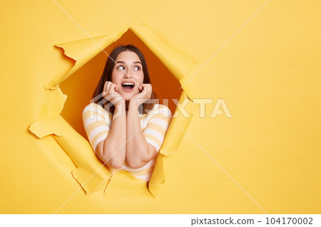 Indoor shot of surprised amazed brunette woman stands in torn paper hole, looks away with open mouth, keeps hands under chin, looking through breakthrough of yellow background. Indoor shot of surprised amazed brunette woman stands in torn paper hole, looks away with open mouth, keeps hands under chin, looking through breakthrough of yellow background. 104170002
