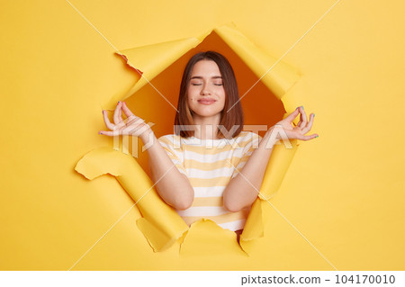 Portrait of relaxed woman wearing striped t shirt looking through breakthrough of yellow background, trying to calm down, relaxing. keeping eyes closed. Portrait of relaxed woman wearing striped t shirt looking through breakthrough of yellow background, trying to calm down, relaxing. keeping eyes closed. 104170010