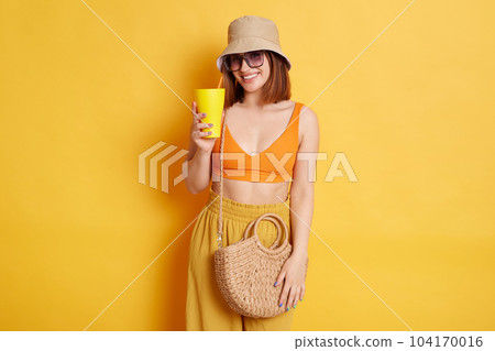 Portrait of delighted young dark haired woman in sunglasses wearing summer clothing posing isolated over yellow background, holding cup with tasty fresh beverage, looking smiling at camera. Portrait of delighted young dark haired woman in sunglasses wearing summer clothing posing isolated over yellow background, holding cup with tasty fresh beverage, looking smiling at camera. 104170016