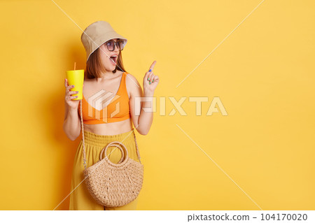 Indoor shot of astonished amazed woman in sunglasses wearing summer clothing posing isolated over yellow background, standing with fresh in hadns and pointing aside and advertising area. 104170020
