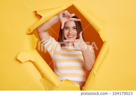 Portrait of smiling winsome woman photographer wearing striped T-shirt breaking through hole in yellow paper, looking through frame from fingers, making focus. 104170023