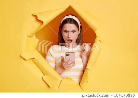 Horizontal shot of surprised shoked woman wearing striped t shirt posing standing through yellow paper torn hole, using smart phone, looking at display with widely opened mouth. 104170050