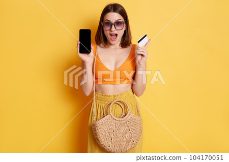 Online shopping. Portrait of shocked woman wearing summer clothing with straw bag standing isolated over yellow background, showing cell phone with empty display, paying for purchase with bank app. 104170051