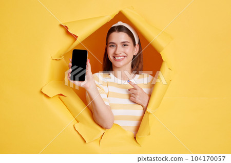 Portrait of smiling positive woman wearing striped t shirt showing cell phone with empty display for advertisement or promotion, posing standing through yellow paper torn hole. 104170057