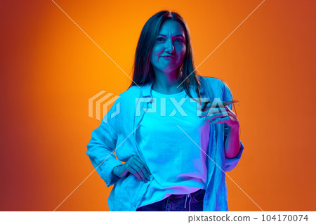 Portrait of young woman in casual clothes posing against gradient orange studio background in neon light Portrait of young woman in casual clothes posing against gradient orange studio background in neon light 104170074