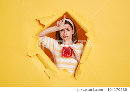 Horizontal shot of sad Caucasian woman wearing hair band and striped t shirt standing in yellow paper hole, holding in hand red alarm clock and showing looser gesture. Horizontal shot of sad Caucasian woman wearing hair band and striped t shirt standing in yellow paper hole, holding in hand red alarm clock and showing looser gesture. 104170286