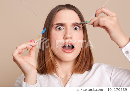 Portrait of shocked scared female in beauty salon holding eyebrow brush and tweezers, looking at camera with big eyes, being shocked after plucking eyebrow procedure. Portrait of shocked scared female in beauty salon holding eyebrow brush and tweezers, looking at camera with big eyes, being shocked after plucking eyebrow procedure. 104170289