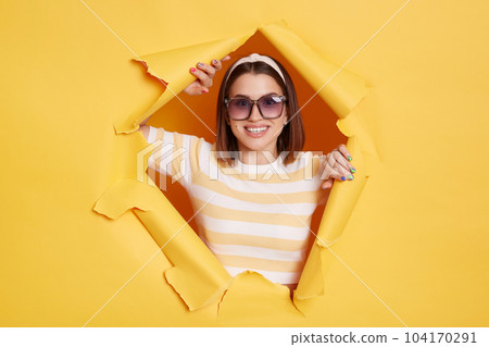 Portrait of cheerful winsome woman wearing striped T-shirt, hair band and sunglasses, breaks through yellow paper background in ripped hole, expressing happiness, looking at camera. 104170291