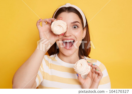Caucasian excited woman holds marshmallows in hands, wearing striped shirt and glasses, covering her eye with sugary sweets, looking at camera with open mouth, posing isolated over yellow background. Caucasian excited woman holds marshmallows in hands, wearing striped shirt and glasses, covering her eye with sugary sweets, looking at camera with open mouth, posing isolated over yellow background. 104170327