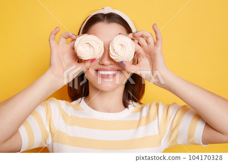 Portrait of smiling happy satisfied woman wearing t shirt and hair band posing isolated on yellow background, standing, having fun, covering eyes with tasty delicious marshmallows. 104170328