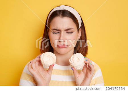 Sad upset woman wearing t shirt and hair band posing isolated on yellow background, being on diet, but wants to eat sugary marshmallows, looking at dessert with pout lips and expressing sorrow. 104170334