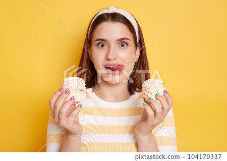 Portrait of funny hungry woman wearing t shirt and hair band posing isolated on yellow background, female being on diet, wants to lick tasty sweets, showing tongue out. Portrait of funny hungry woman wearing t shirt and hair band posing isolated on yellow background, female being on diet, wants to lick tasty sweets, showing tongue out. 104170337