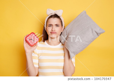 Indoor shot of sad woman with alarm clock awakening hard early wearing casual striped t-shirt posing isolated on yellow background, Caucasian female has no enough sleep, want more rest at weekends. Indoor shot of sad woman with alarm clock awakening hard early wearing casual striped t-shirt posing isolated on yellow background, Caucasian female has no enough sleep, want more rest at weekends. 104170347