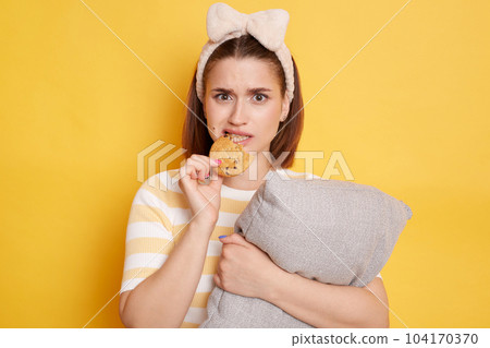 Portrait of young adult dark haired woman biting cookie eating sweets isolated on yellow background, being in bad mood, looking at camera with sad expression. Portrait of young adult dark haired woman biting cookie eating sweets isolated on yellow background, being in bad mood, looking at camera with sad expression. 104170370
