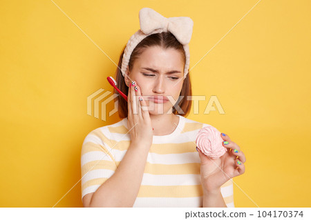 Portrait of sad sick Caucasian woman wearing t shirt and hair band, holding marshmallow and toothbrush, having dental problems after drinking sweets, posing isolated on yellow background. Portrait of sad sick Caucasian woman wearing t shirt and hair band, holding marshmallow and toothbrush, having dental problems after drinking sweets, posing isolated on yellow background. 104170374