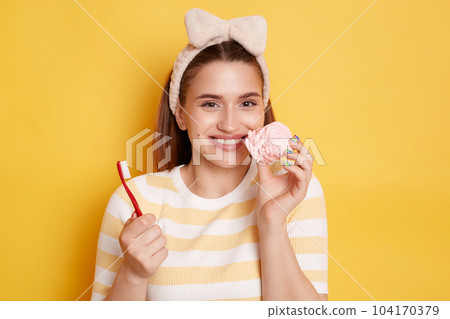 Horizontal shot of smiling satisfied dark haired woman wearing striped shirt and hair band posing isolated over yellow background, holding toothbrush and tasty marshmallow, has not problems with teeth Horizontal shot of smiling satisfied dark haired woman wearing striped shirt and hair band posing isolated over yellow background, holding toothbrush and tasty marshmallow, has not problems with teeth 104170379