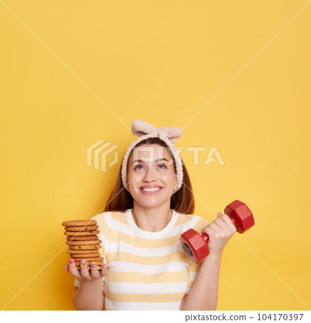Portrait of smiling woman wearing striped shirt and hair band holding red dumbbell and cookies, posing isolated over yellow background, looking up at copy space for advertisement or promotion. 104170397