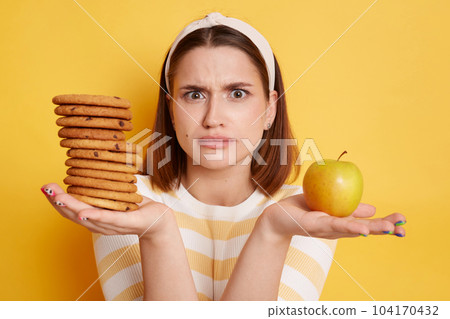 Portrait of confused woman wearing striped shirt and hair band, looking at camera with puzzlement, holding cookies and apple, does not know what to choose, posing isolated over yellow background. 104170432
