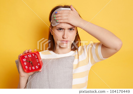 Portrait of sad upset tired woman wearing striped t shirt and blindfold standing with alarm clock and pillow in hands isolated over yellow background, fells sleepy and having headache. 104170449