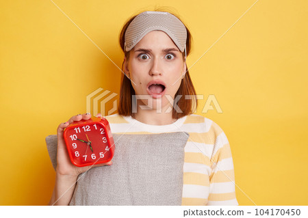 Shocked surprised woman wearing striped shirt and sleeping mask, holding pillow and red alarm clock, posing isolated over yellow background, looking at camera with open mouth and big eyes, being late. 104170450