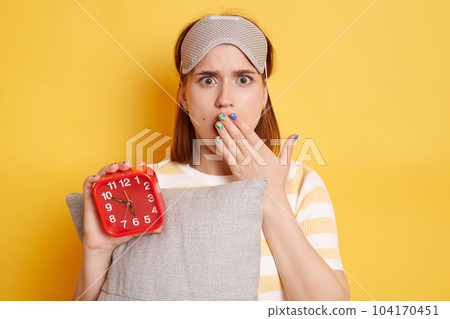 Shocked woman wearing striped t shirt and blindfold standing with alarm clock and pillow in hands isolated over yellow background, looking at camera with big eyes, covering mouth with palm. 104170451