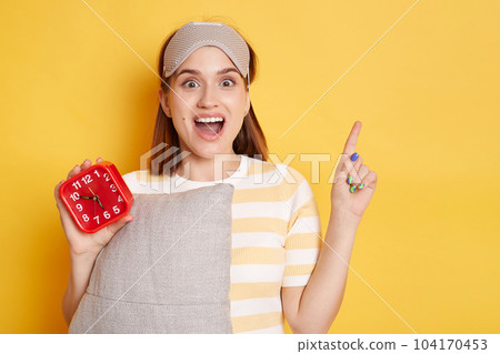 Horizontal shot of excited Caucasian woman looking at camera prepares for sleep holds soft pillow dressed in sleepmask on forehead, pointing aside with finger, showing copy space for advertisement. Horizontal shot of excited Caucasian woman looking at camera prepares for sleep holds soft pillow dressed in sleepmask on forehead, pointing aside with finger, showing copy space for advertisement. 104170453