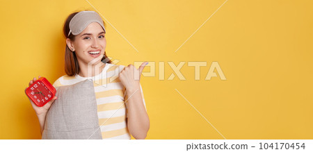 Portrait of smiling young Caucasian woman wearing striped t shirt and blindfold on her head posing isolated over yellow background, holding alarm clock and pillow, pointing away at copy space. 104170454
