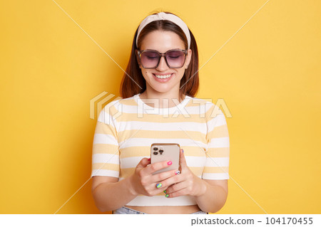Indoor shot of smiling delighted woman wearing sunglasses, striped T-shirt and hair band posing isolated over yellow background, holding smart phone, browsing Internet, checking social networks. Indoor shot of smiling delighted woman wearing sunglasses, striped T-shirt and hair band posing isolated over yellow background, holding smart phone, browsing Internet, checking social networks. 104170455