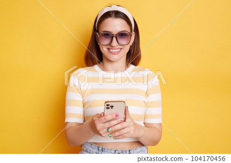 Indoor shot of attractive young woman wearing striped t-shirt standing with smart phone in hands, using cell phone, looking at camera with smile, posing isolated over yellow background. 104170456