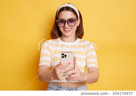 Indoor shot of smiling satisfied woman wearing striped t shirt and jeans, holding smart phone in hands, surfing internet, checking social networks, posing isolated over yellow background. 104170457