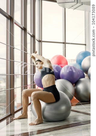 Fitness girl working out at gym with big window. Girl wearing black leggins and sport top. Young woman doing excercises with a fitness ball. 104173089