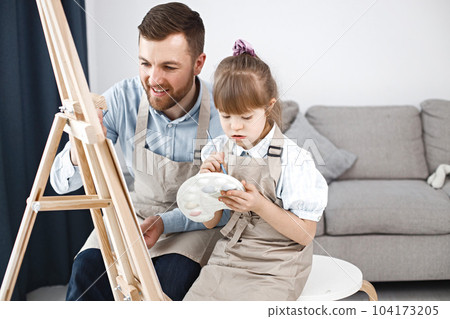 Little girl with Down syndrome wearing white shirt and beige apron. Girl with her father is painting on easel together. Father helping his daughter to paint. Little girl with Down syndrome wearing white shirt and beige apron. Girl with her father is painting on easel together. Father helping his daughter to paint. 104173205