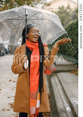 Young black woman with a long locs hairstyles standing outside with a transparent umbrella. Woman wearing brown coat, orange scarf and black hat. Woman in glasses resting outside on autumn. 104173283