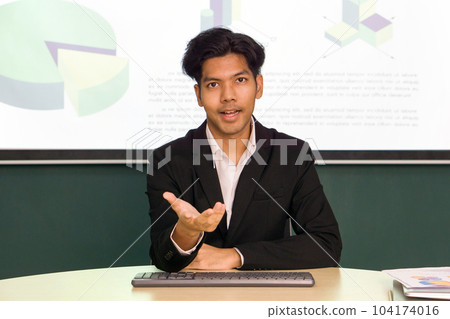 Young asian businessman in white shirt and black suit looking at camera, recording self-presentation video or sharing professional skill. Multicolor chart are on presentation monitor in the background Young asian businessman in white shirt and black suit looking at camera, recording self-presentation video or sharing professional skill. Multicolor chart are on presentation monitor in the background 104174016