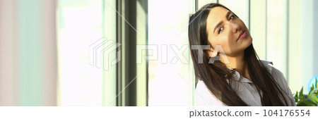 Dreamy young business woman is sitting on windowsill near huge window in modern office sun is shining brightly Dreamy young business woman is sitting on windowsill near huge window in modern office sun is shining brightly 104176554