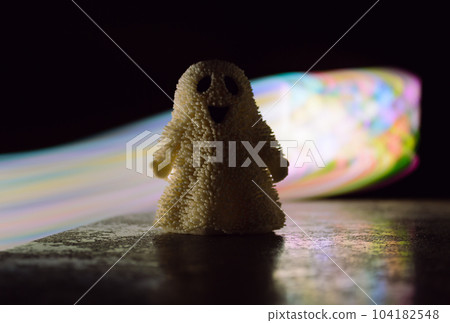 A toy ghost stands in the semi-darkness against the background of a beam of light in the dark A toy ghost stands in the semi-darkness against the background of a beam of light in the dark 104182548