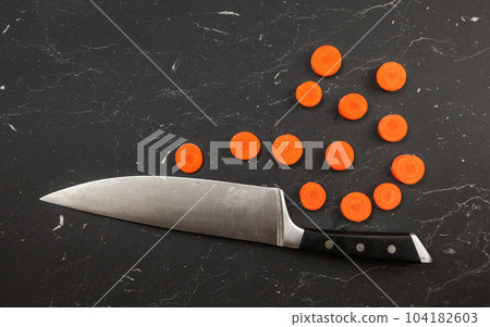 Top down view, chefs knife and small circles of sliced carrot on black marble like working board. Top down view, chefs knife and small circles of sliced carrot on black marble like working board. 104182603