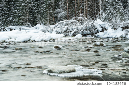 Small creek in winter, snow covered trees and shore background, ice on top of rocks at river 104182656