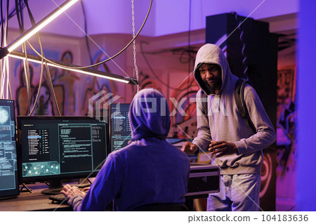 African american hacker bringing black box with technology for cyberattack. Young man in hood showing case with tool for data breach and explaining internet crime plan in hideout place 104183636